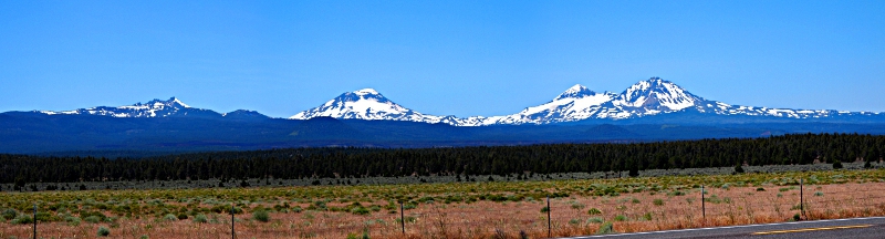 [Several photos stitched together - The Three Sisters are three mountains heavily (but not completely) covered in snow. Two of the 'Sisters' are nearly atop each other while the third sister is a short distance to the left. Even further to the left is Broken Top which is much smaller and less snow covered than the other three.]